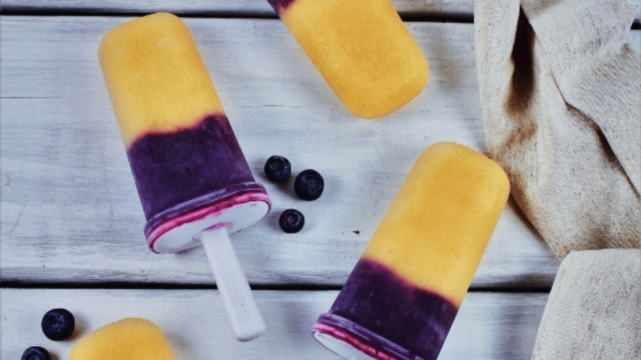 Popsicle maison 2 couleurs, mauve dans le bas et orange dans le haut sur une table en bois blanche et quelques bleuets frais