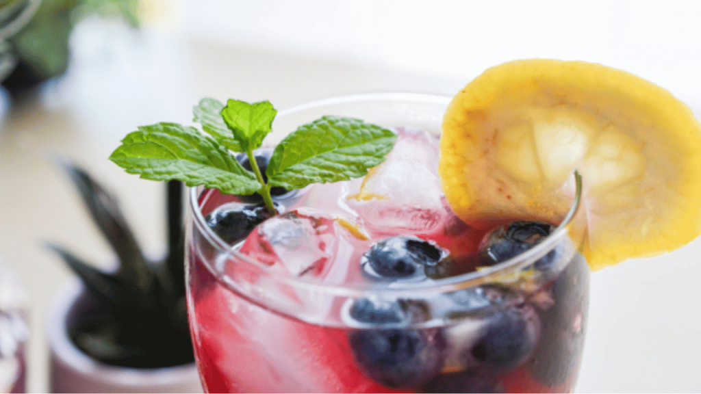 Gros plan sur un verre de cocktail roure framboise décoré d'une tranche de citron, des bleuets et une feuille de menthe