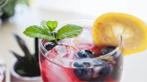 Gros plan sur un verre de cocktail roure framboise décoré d'une tranche de citron, des bleuets et une feuille de menthe