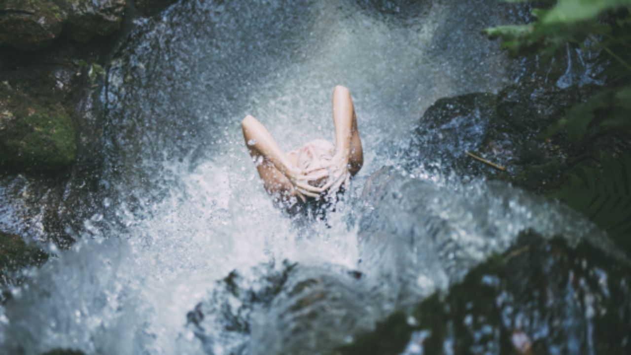 Personne se tenant sous une petite cascade en nature, les bras levés, tandis que l’eau éclabousse autour d’elle
